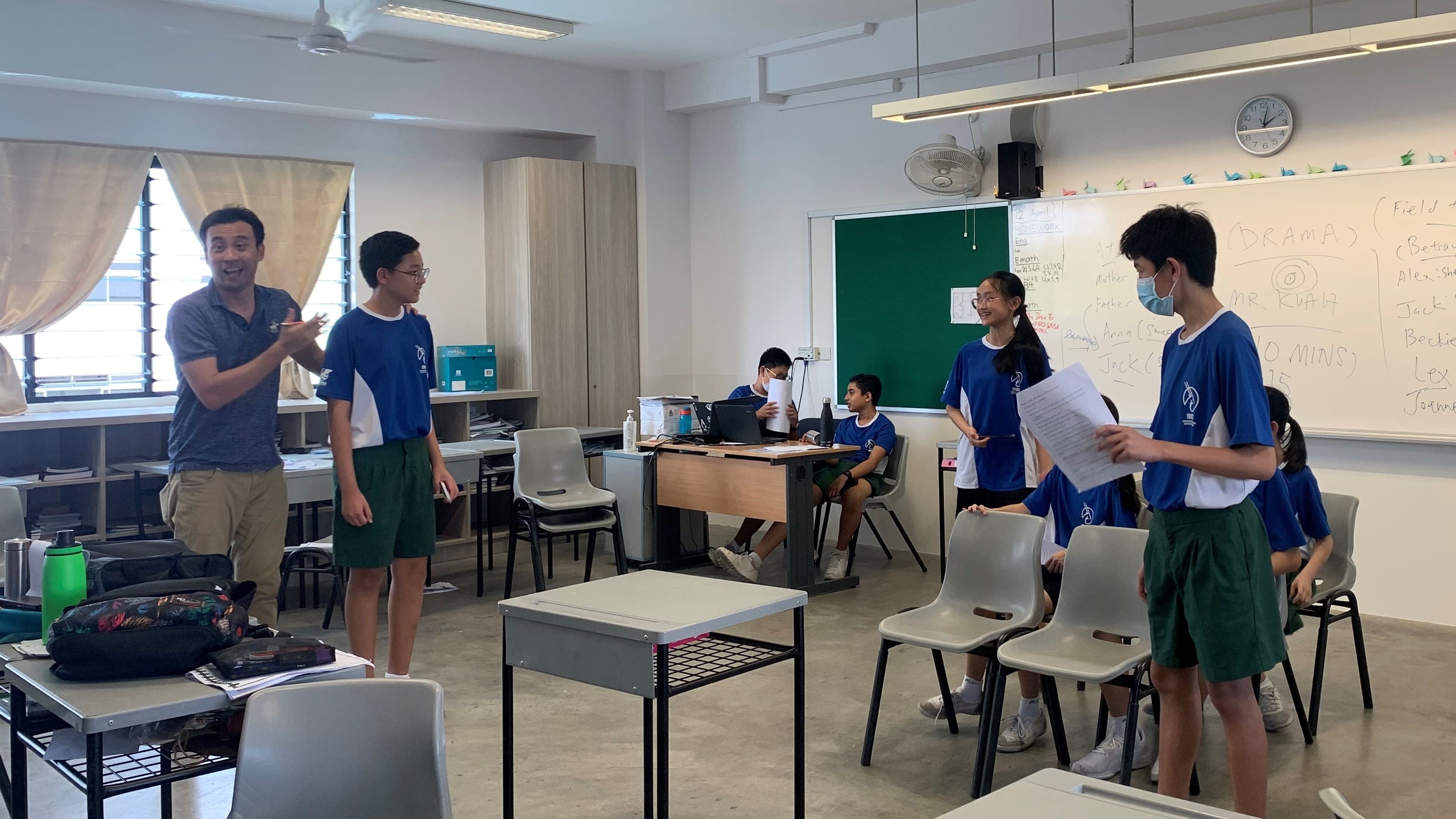 A teacher directs students in blue uniforms during a drama class in a classroom with a whiteboard.